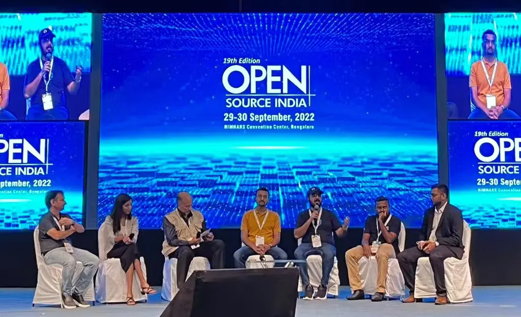 Joshua Poddoku, far right in a dark suit, on a seven-person panel at Open Source India 2022, under the conference screens at NIMHANS Convention Centre, Bengaluru